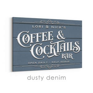 May include: A dusty denim-colored sign with white lettering that reads "LORI & NICK'S COFFEE & COCKTAILS BAR." The sign has a rustic, wooden plank design and decorative flourishes. The text "OPEN DAILY - SELF SERVE" is also visible.