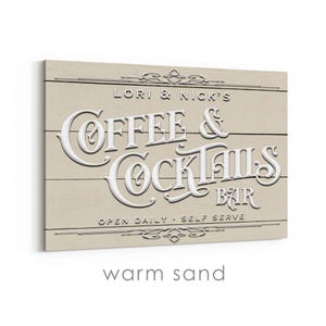 May include: A rectangular wooden sign with the words "Coffee & Cocktails Bar" in white, raised lettering. The sign is beige with horizontal wood grain lines. The text "Lori & Nick's" and "Open Daily - Self Serve" are also visible.
