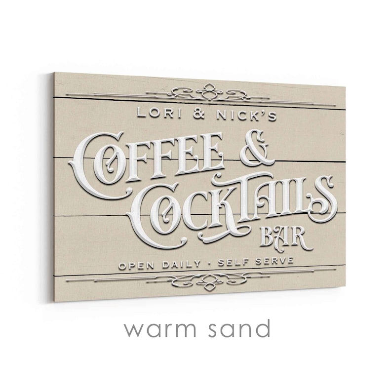 May include: A rectangular wooden sign with the words "Coffee & Cocktails Bar" in white, raised lettering. The sign is beige with horizontal wood grain lines. The text "Lori & Nick's" and "Open Daily - Self Serve" are also visible.