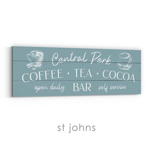 May include: A rectangular, teal wooden sign featuring white text and illustrations. The sign says "Central Perk" with the words "Coffee, Tea, Cocoa, Bar, self service, open daily". The sign has illustrations of coffee cups.