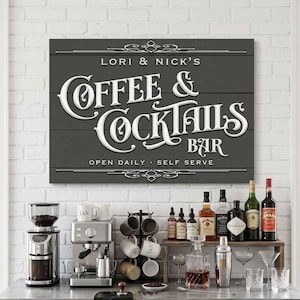 May include: A dark gray sign with white lettering that reads "LORI & NICK'S COFFEE & COCKTAILS BAR." Below the sign is a coffee and cocktail bar setup with a coffee grinder, espresso machine, mugs, and liquor bottles.