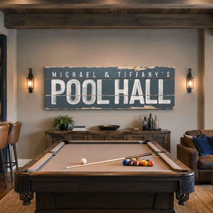 May include: A rectangular wooden wall sign with a dark blue distressed finish and white painted text that reads MICHAEL & TIFFANY'S POOL HALL. The sign is composed of three horizontal planks and is mounted on a wall above a pool table.