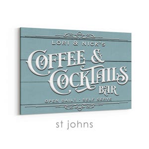 May include: A rectangular sign with the words "LORI & NICK'S COFFEE & COCKTAILS BAR" in white, stylized lettering. The sign is light blue with horizontal wood grain. The text "OPEN DAILY - SELF SERVE" is also visible. The word "st johns" is at the bottom.
