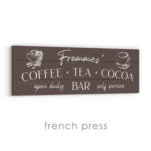 May include: A rectangular wooden sign with a dark brown background. The sign features white text and illustrations related to a coffee, tea, and cocoa bar. The text includes "Frommes'", "COFFEE &bull; TEA &bull; COCOA", "open daily BAR self service".