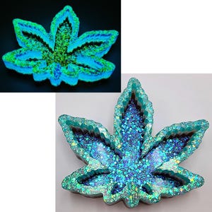 May include: A leaf-shaped tray with a glittery, iridescent finish. The tray is primarily teal and blue, with a dark brown interior. The design is reminiscent of a cannabis leaf. The tray is shown in two different lighting conditions, one with a black background and a glowing effect.