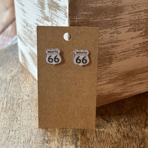 Puede incluir: Un par de pendientes de la Ruta 66 en una tarjeta marrón. Los pendientes son plateados con fondo blanco y texto negro.