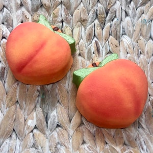 Puede incluir: Dos adornos con forma de melocotón, en tonos naranja y rojo, con hojas verdes y pequeños tallos marrones. Se encuentran sobre una superficie tejida de color marrón claro, creando un ambiente natural y rústico. Los melocottones tienen un aspecto texturizado.