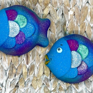 May include: Two blue fish-shaped bath bombs with glitter accents. The fish have a gradient of blue and purple, with silver, purple, and green scales. The fish have a small, open mouth and a white eye. The background is a woven, light brown material.