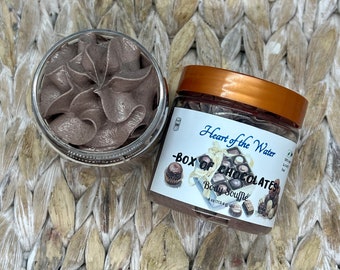 May include: A brown glass jar with a brown whipped body souffle inside. The jar has a gold lid and a label that says "Heart of the Water - Box of Chocolates Body Souffle 4 oz (113.4 g) net wt."