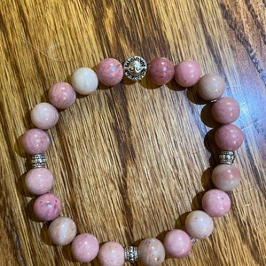May include: A pink and white gemstone bracelet with silver beads and a silver charm. The bracelet is made of round beads and is strung on a stretchy cord.