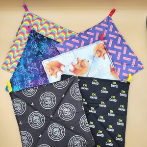 May include: A set of six colorful pot holders. The pot holders are made of fabric and have different patterns, including rainbows, stars, bacon, and pigs. The pot holders are all square and have a loop on the top for hanging.