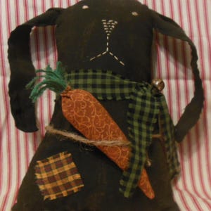 May include: A black fabric bunny with a green and black plaid ribbon, holding a brown carrot. The bunny has a bell on its neck and is made in a primitive style.
