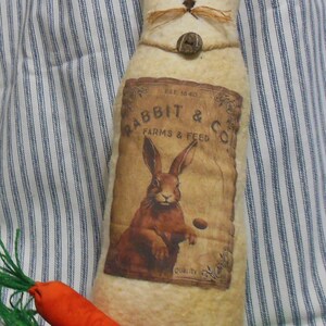 May include: A white felt bunny with a brown felt nose and whiskers. The bunny is wearing a brown fabric collar with a wooden button. The bunny is holding a vintage-style advertisement for "Rabbit & Co. Farms & Feed" featuring a brown rabbit.
