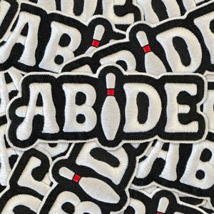 May include: Embroidered patch with the word "ABIDE" in white, outlined in black, with a bowling pin replacing the "I". The bowling pin has a red stripe. The patch is on a black background.