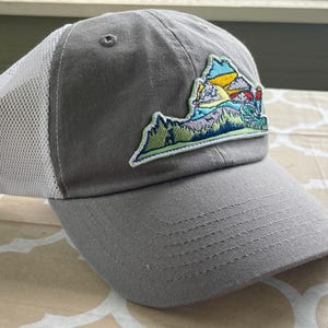 May include: Gray baseball cap with a white mesh back and a curved brim. The cap features an embroidered patch of a colorful landscape with mountains, trees, and a sunset. The hat is a casual accessory.