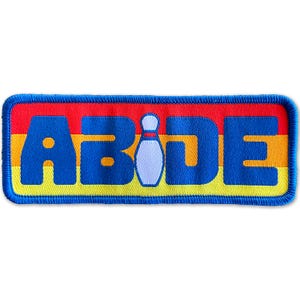 May include: A blue and yellow embroidered patch with the word "ABIDE" and a white bowling pin in the center. The patch has a red, orange, and yellow striped background.