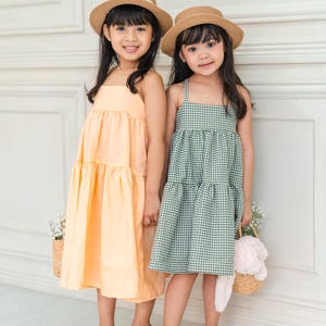 May include: Two girls wearing straw hats and summer dresses. One girl is wearing a light orange tiered dress and the other is wearing a green and white gingham tiered dress.