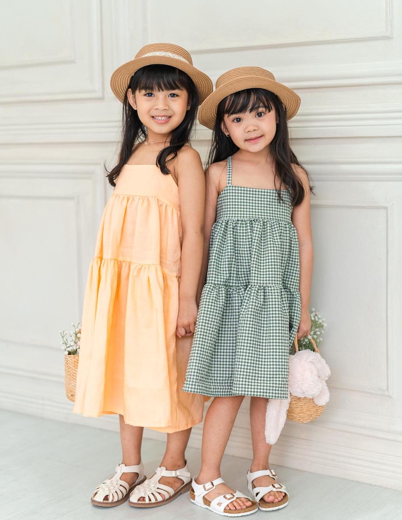 May include: Two girls wearing straw hats and summer dresses. One girl is wearing a light orange tiered dress and the other is wearing a green and white gingham tiered dress.