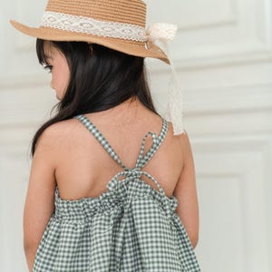 May include: A young girl wearing a green and white gingham dress with a brown straw hat with a white lace trim. The dress has a tie at the back.