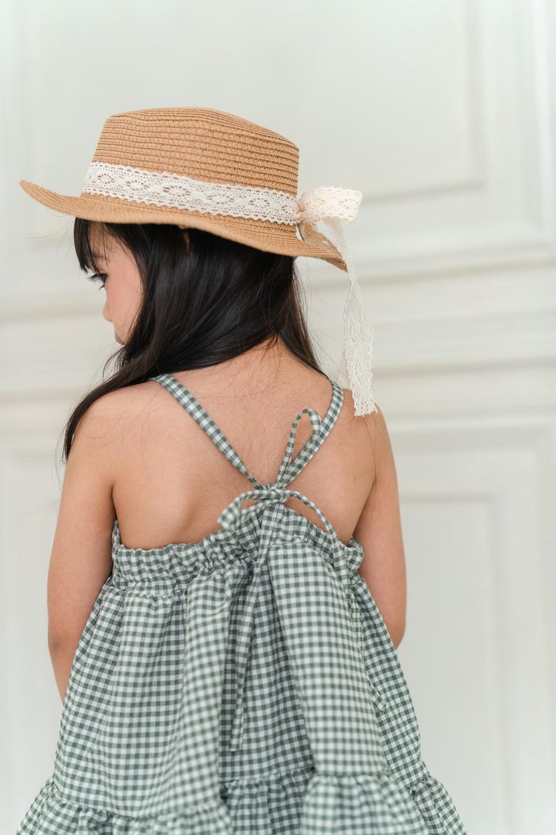 May include: A young girl wearing a green and white gingham dress with a brown straw hat with a white lace trim. The dress has a tie at the back.