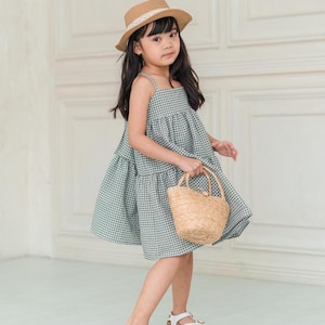 May include: A young girl wearing a green and white gingham dress, a straw hat with a white ribbon, and white sandals. She is holding a small woven basket.