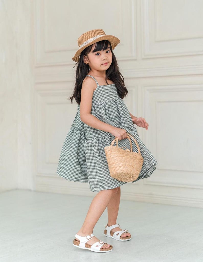 May include: A young girl wearing a green and white gingham dress, a straw hat with a white ribbon, and white sandals. She is holding a small woven basket.