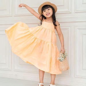 May include: A young girl wearing a light yellow tiered dress and a straw hat, holding a small wicker basket with white flowers. She is smiling and standing in front of a white wall.