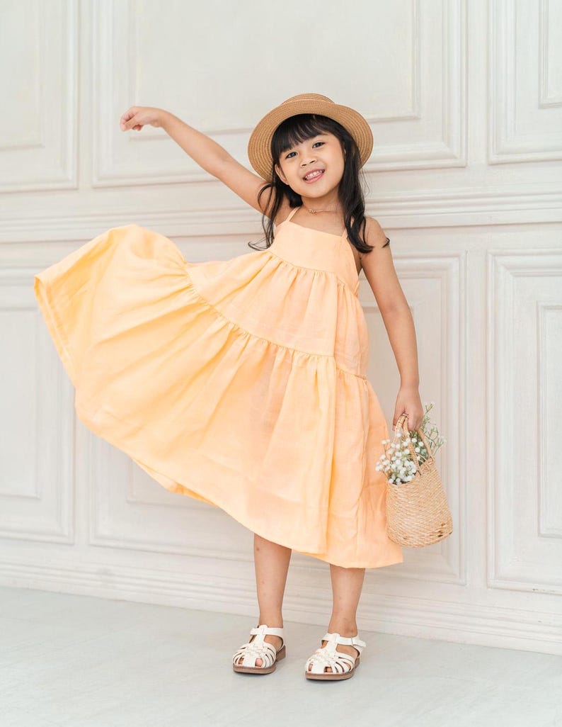May include: A young girl wearing a light yellow tiered dress and a straw hat, holding a small wicker basket with white flowers. She is smiling and standing in front of a white wall.