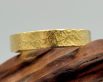 Puede incluir: Un anillo de oro con una banda plana y un acabado de superficie texturizado y martillado. El anillo presenta un ancho constante en toda su circunferencia, mostrando un color amarillo metálico con intrincadas hendiduras irregulares.
