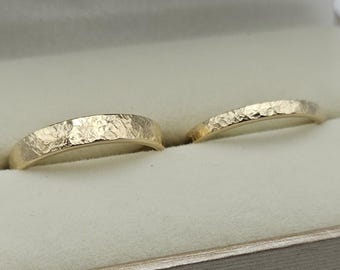 May include: Two gold rings with a textured, hammered finish. The bands have a distinct, uneven surface that catches the light, creating a rugged appearance. The rings are positioned side by side, showcasing their metallic luster and craftsmanship.
