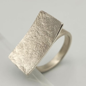 Op de afbeelding: Een zilveren ring met een rechthoekige, getextureerde voorkant. De band van de ring is glad en afgerond, wat contrasteert met het ruwe oppervlak van het rechthoekige element. De ring staat tegen een witte achtergrond.