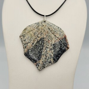 May include: A black cord necklace with a large, irregularly shaped pendant made of polished stone. The stone is a mix of black, white, and brown with a speckled pattern.