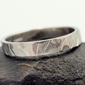 May include: A silver-toned metal ring featuring a textured outer surface with a series of irregular, etched, diagonal, and horizontal lines that create a faceted appearance across the band, showcasing a metallic sheen and a polished finish.