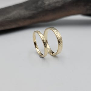 Puede incluir: Dos anillos de oro con un acabado texturizado y martillado en la superficie exterior. Los anillos son circulares, con una banda notablemente m&aacute;s delgada que la otra. Ambos anillos presentan un brillo met&aacute;lico reflectante y color oro amarillo.