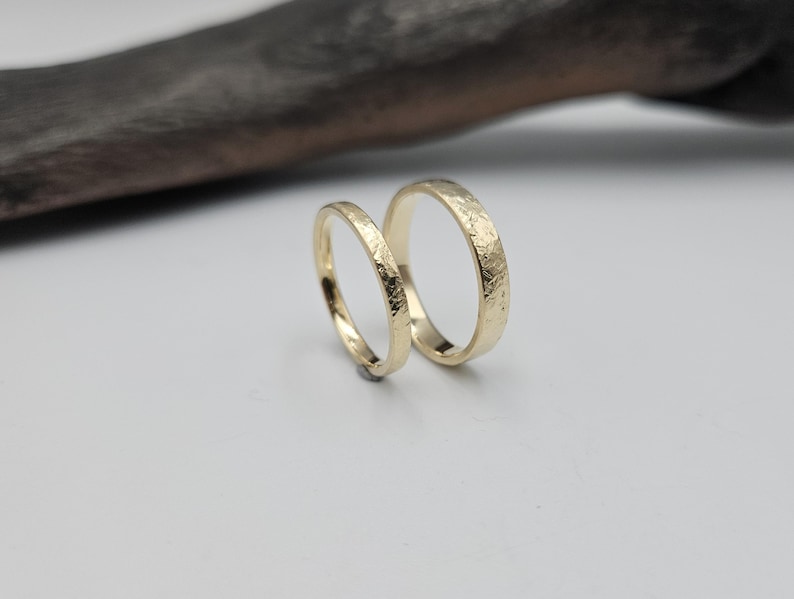 Puede incluir: Dos anillos de oro con un acabado texturizado y martillado en la superficie exterior. Los anillos son circulares, con una banda notablemente m&aacute;s delgada que la otra. Ambos anillos presentan un brillo met&aacute;lico reflectante y color oro amarillo.
