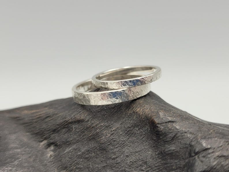 May include: Two silver-colored rings with a textured, matte finish. The rings have a flat profile and a metallic sheen, showing a cross-hatched, scratched surface pattern across the outer bands. The rings are stacked slightly offset from each other.