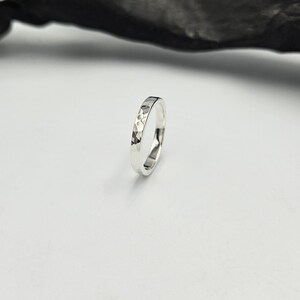 May include: A silver ring with a hammered, faceted texture on the outer surface. The band has a polished, reflective finish that catches the light, creating a series of small, irregular geometric indentations around the entire circumference.