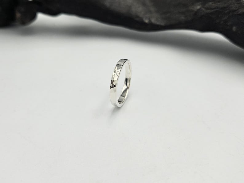 May include: A silver ring with a hammered, faceted texture on the outer surface. The band has a polished, reflective finish that catches the light, creating a series of small, irregular geometric indentations around the entire circumference.