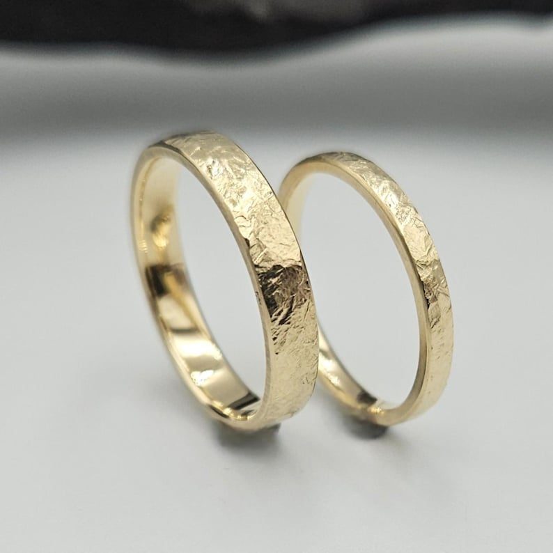 Puede incluir: Dos anillos de oro con un acabado texturizado y martillado en la superficie exterior. Los anillos tienen un perfil plano y un interior pulido. El metal tiene un tono amarillo c&aacute;lido y la superficie presenta hendiduras irregulares y reflectantes.