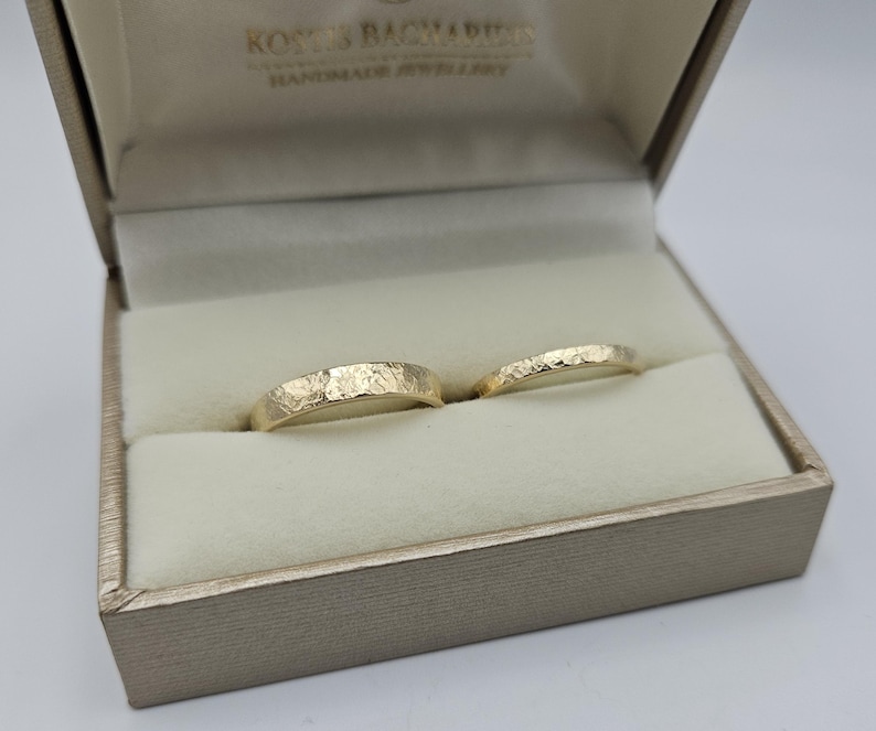 Puede incluir: Dos anillos de oro con un acabado de superficie texturizado y martillado, presentados dentro de un joyero beige. El interior de la tapa de la caja presenta el texto KOSTIS BACHARDIS HANDMADE JEWELLERY impreso en color oscuro sobre la tela.