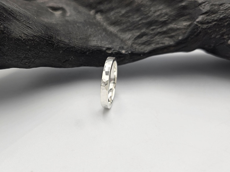 May include: A silver ring with a hammered, faceted texture on the outer surface. The band has a polished, reflective finish and a consistent width, showcasing a series of small, flat, geometric indentations that create a light-catching effect.