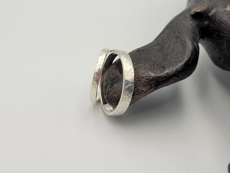 May include: Two silver rings with a textured, matte finish. The bands have a consistent width and a slightly irregular, hammered surface appearance. The rings are positioned together, showing their circular shape and metallic silver color.