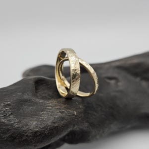 Puede incluir: Dos anillos de oro entrelazados con una superficie texturizada y martillada. Los anillos son de forma circular con un tono amarillo met&aacute;lico, mostrando un contraste claro entre la textura exterior rugosa e irregular y el interior liso y pulido.