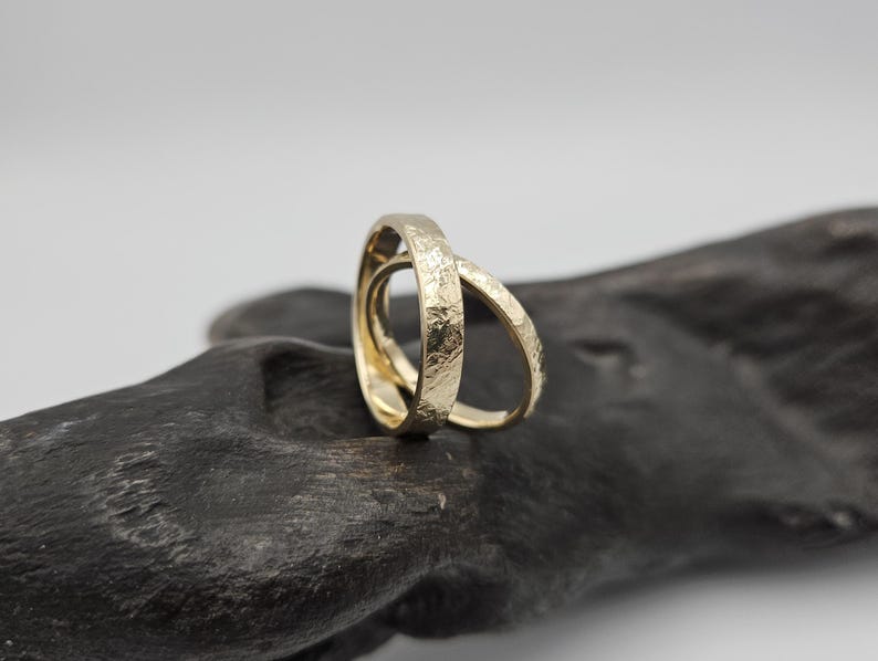 Puede incluir: Dos anillos de oro entrelazados con una superficie texturizada y martillada. Los anillos son de forma circular con un tono amarillo met&aacute;lico, mostrando un contraste claro entre la textura exterior rugosa e irregular y el interior liso y pulido.