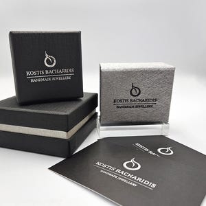 Puede incluir: Dos cajas de joyer&iacute;a cuadradas negras con superficies texturizadas, una apilada sobre la otra, y una caja texturizada gris plateado. Cada caja presenta el logotipo y el texto blanco KOSTIS BACHARIDIS HANDMADE JEWELLERY. Se incluyen dos tarjetas negras con el mismo logotipo.
