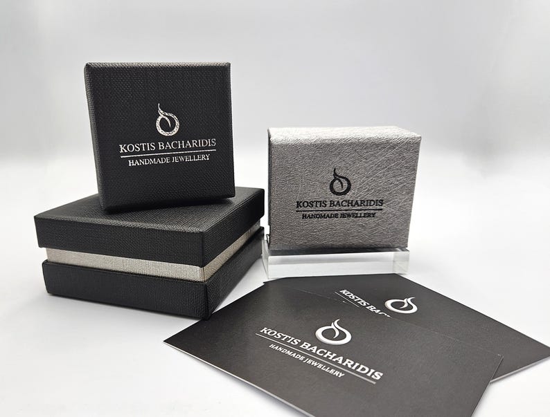 Puede incluir: Dos cajas de joyer&iacute;a cuadradas negras con superficies texturizadas, una apilada sobre la otra, y una caja texturizada gris plateado. Cada caja presenta el logotipo y el texto blanco KOSTIS BACHARIDIS HANDMADE JEWELLERY. Se incluyen dos tarjetas negras con el mismo logotipo.