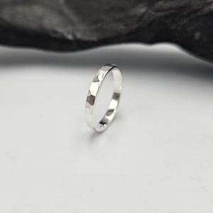 May include: A silver ring with a hammered, faceted texture on the outer surface. The band has a polished, reflective finish that catches the light, creating distinct highlights across the multiple flat, angled planes of the metal surface.