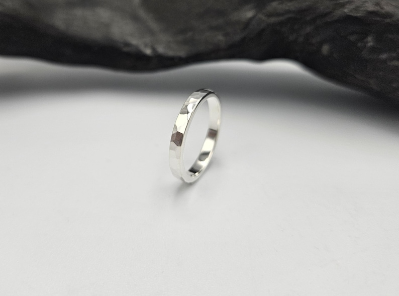 May include: A silver ring with a hammered, faceted texture on the outer surface. The band has a polished, reflective finish that catches the light, creating distinct highlights across the multiple flat, angled planes of the metal surface.