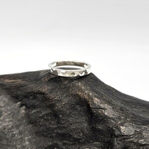 May include: A silver-toned ring featuring a hammered, faceted texture across its outer surface. The band has a consistent width and a reflective metallic finish, showcasing multiple small, irregular indentations that create a light-catching effect.