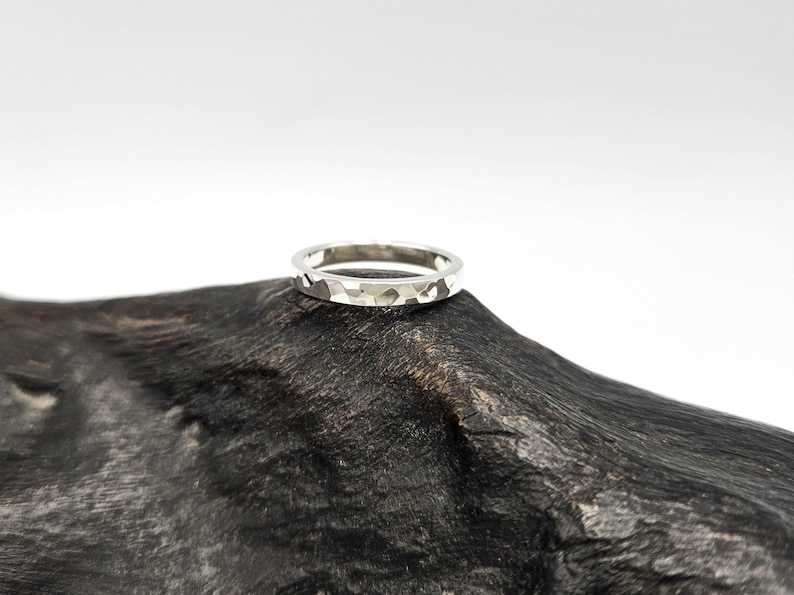 May include: A silver-toned ring featuring a hammered, faceted texture across its outer surface. The band has a consistent width and a reflective metallic finish, showcasing multiple small, irregular indentations that create a light-catching effect.